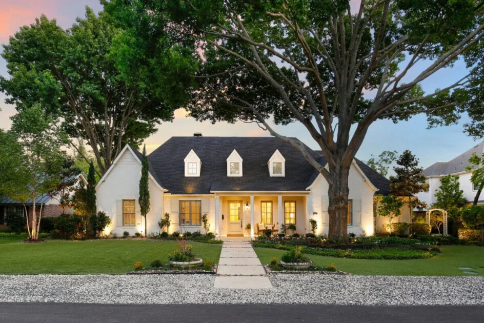 White farmhouse with dark roof, dormers, wood portico, concrete walkway, and landscape lighting